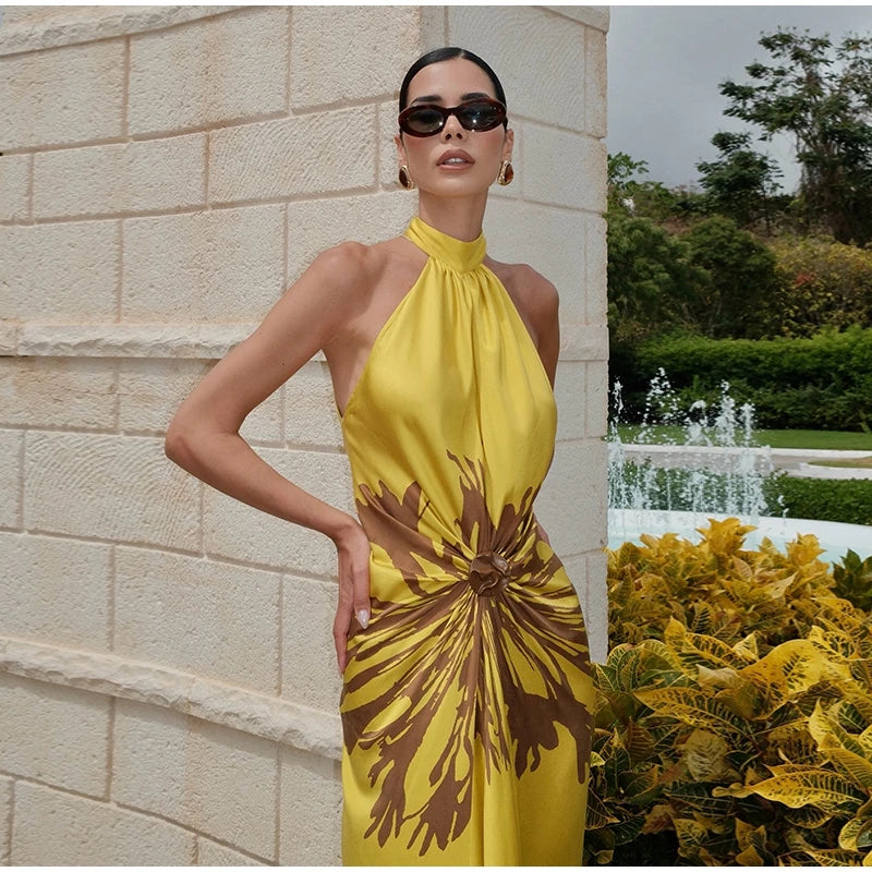 A woman wearing a bright yellow halter-neck dress with a large brown abstract floral print, standing outdoors next to a light stone wall and greenery.
