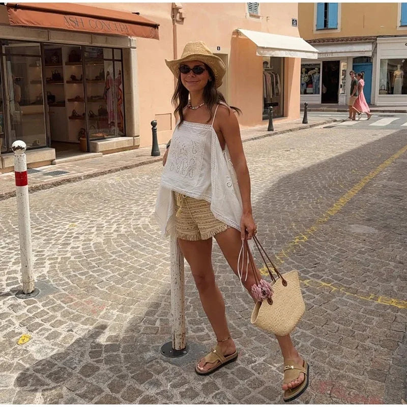 A woman stands on a cobblestone street wearing a white embroidered camisole,