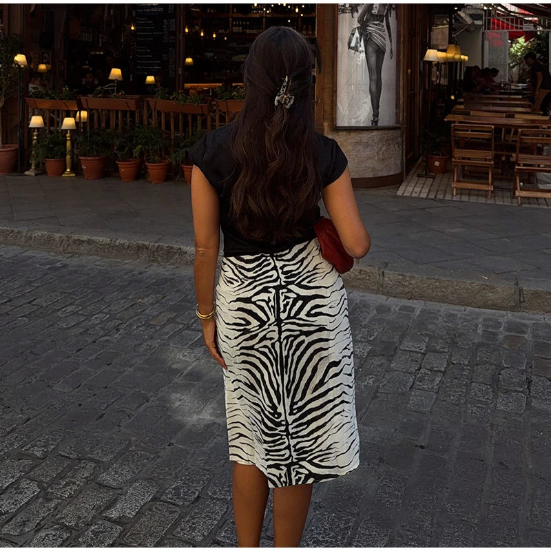 A woman viewed from behind wearing a black short-sleeve top and a black-and-white zebra print midi skirt, standing on a cobblestone street in front of a restaurant.