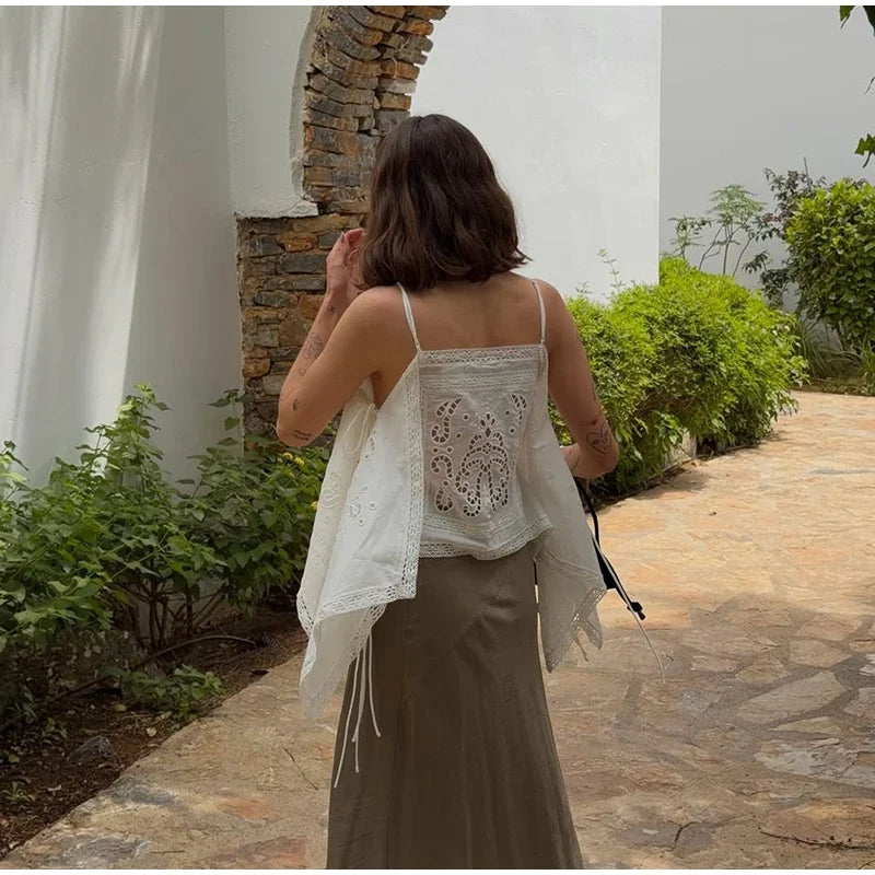 A woman viewed from behind wearing a white embroidered lace camisole top with thin straps and a sheer draped overlay,