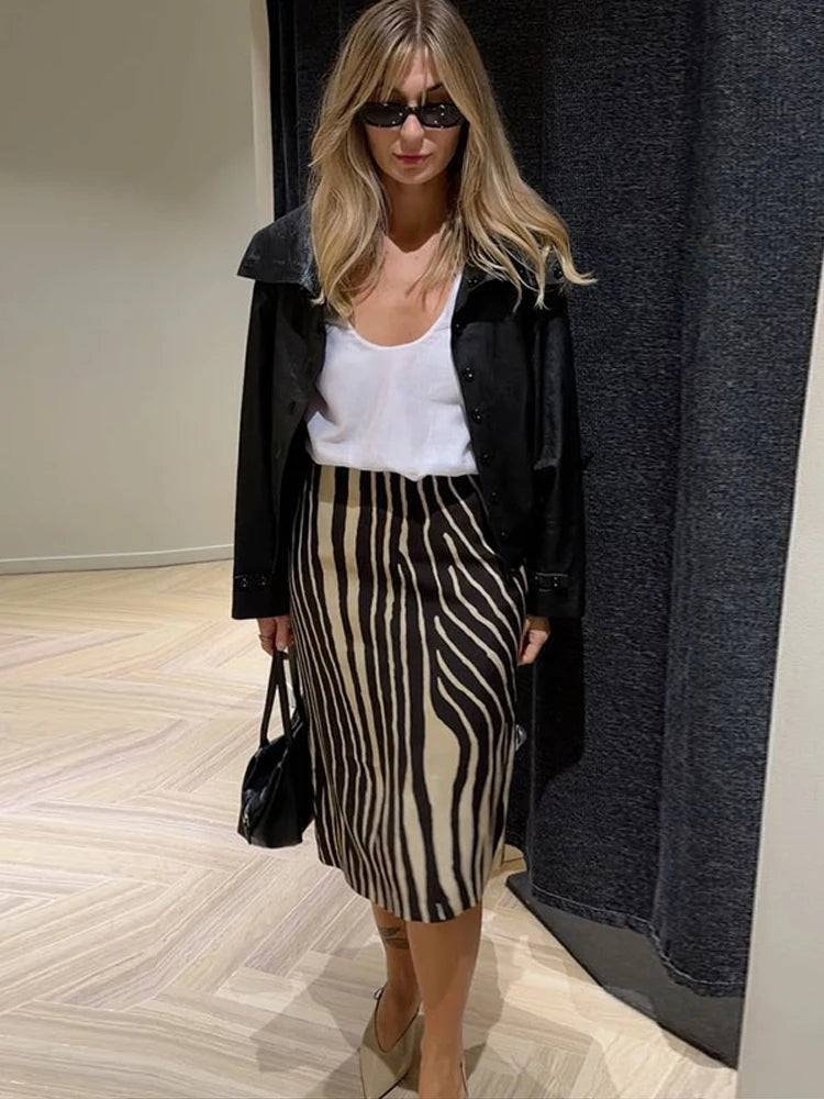 A woman modeling a black and beige zebra-print midi skirt, styled with a white tank top, black jacket, black sunglasses, and beige heels.