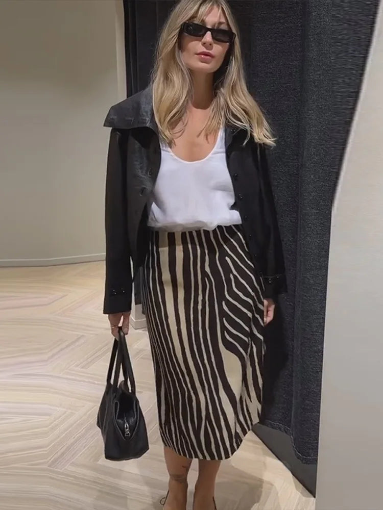 A woman wearing a black leather jacket, white tank top, and a black and beige zebra-print midi skirt, holding a black handbag. She is wearing black sunglasses and standing in an indoor setting with a light wood floor and a dark gray curtain.