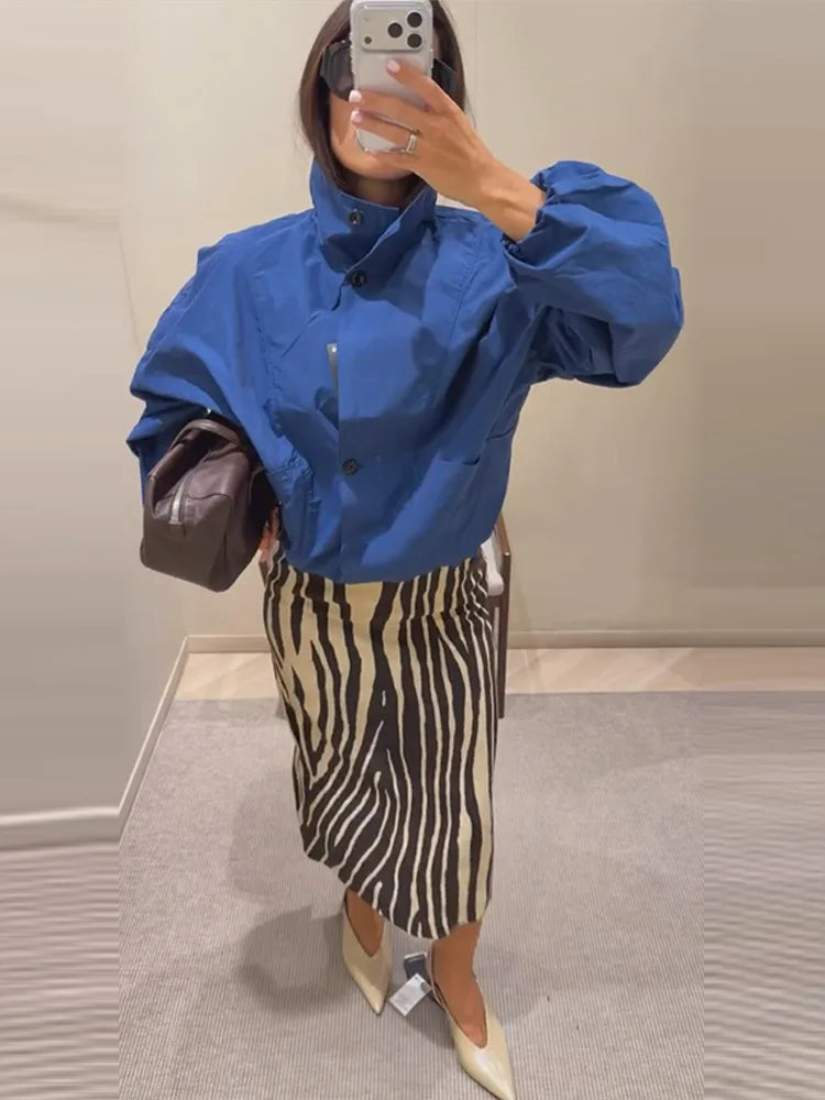 A woman takes a mirror selfie in a clothing store showing a blue long-sleeve button-up shirt with a high collar and a beige and black zebra-print midi skirt.