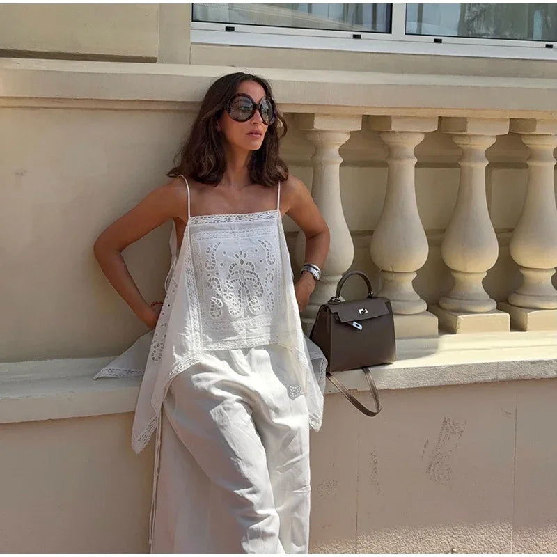 A woman wearing a white embroidered camisole top and matching white wide-leg pants, leaning against a beige stone balustrade wall. She is wearing dark sunglasses and holding a dark brown structured handbag.