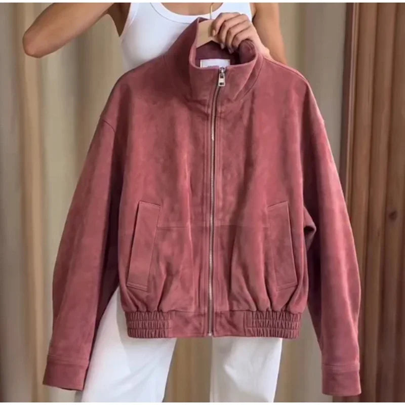 A dusty rose pink suede zip-up jacket with a stand-up collar, elasticized hem and cuffs, and front flap pockets, held up by a person wearing a white tank top and white pants.